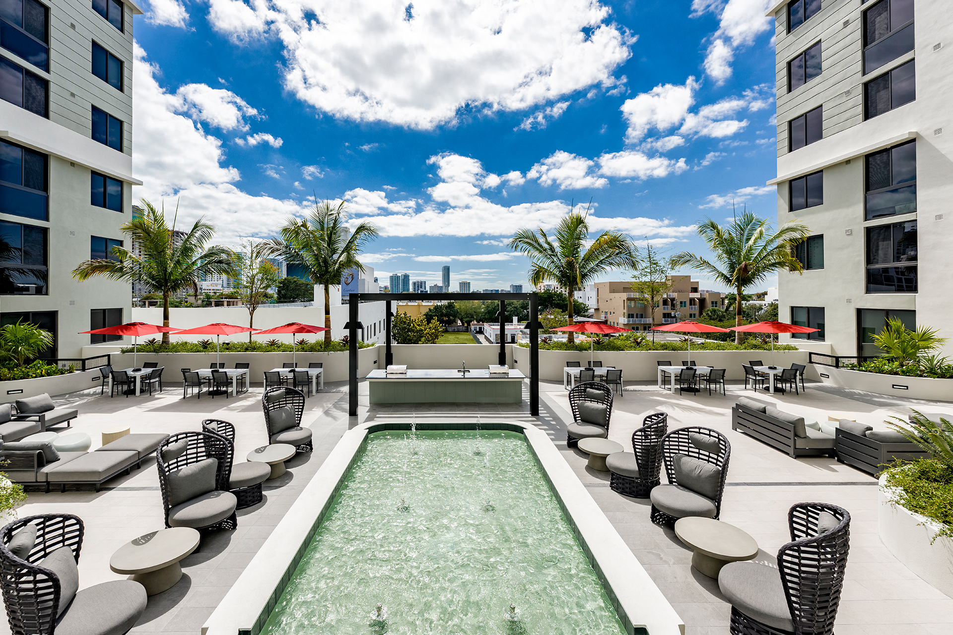 Two-Bedroom Apartments in Little Havana, FL - Oasis Liberty Square - Rooftop Courtyard With Water Feature, Lounge Chairs, Red Umbrellas, Palm Trees, and Open Skyline Views Between Modern Buildings.
